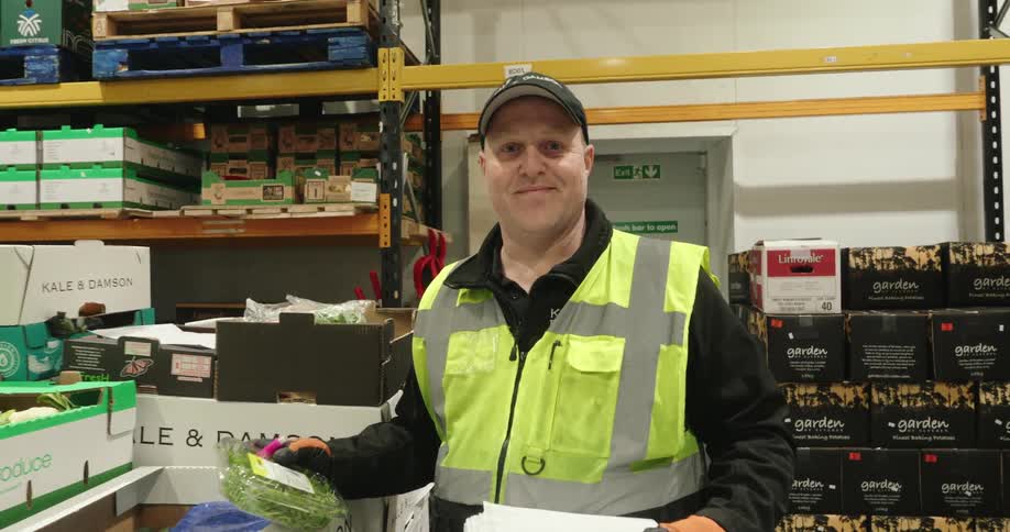 A warehouse worker smiles at the camera while holding produce in a storage area filled with boxes