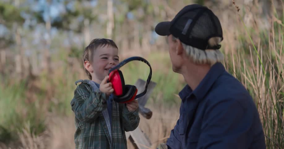 A young boy wearing earmuffs playfully interacts with his father outdoors in a grassy field