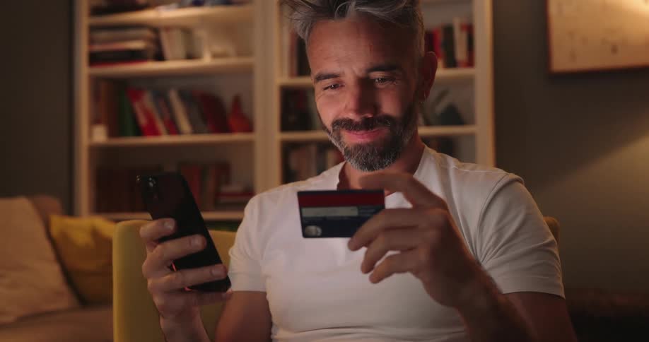 A man with salt and pepper hair and beard sits in a chair holding a credit card and looking at his smartphone with a smile