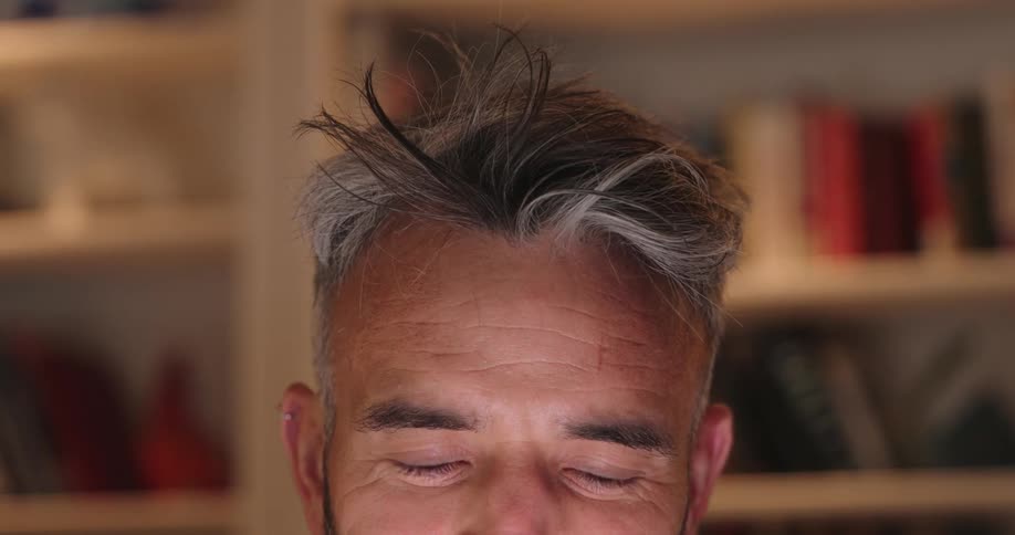 A bearded man with white hair looks at a screen indoors as its light reflects on his face