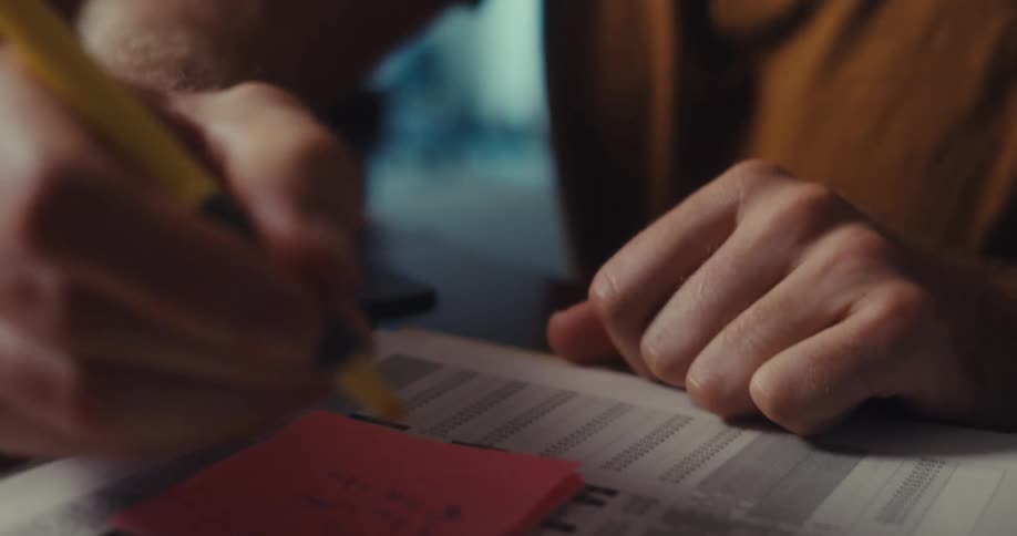 A man is intently reviewing a printed document and highlighting information while working on a laptop