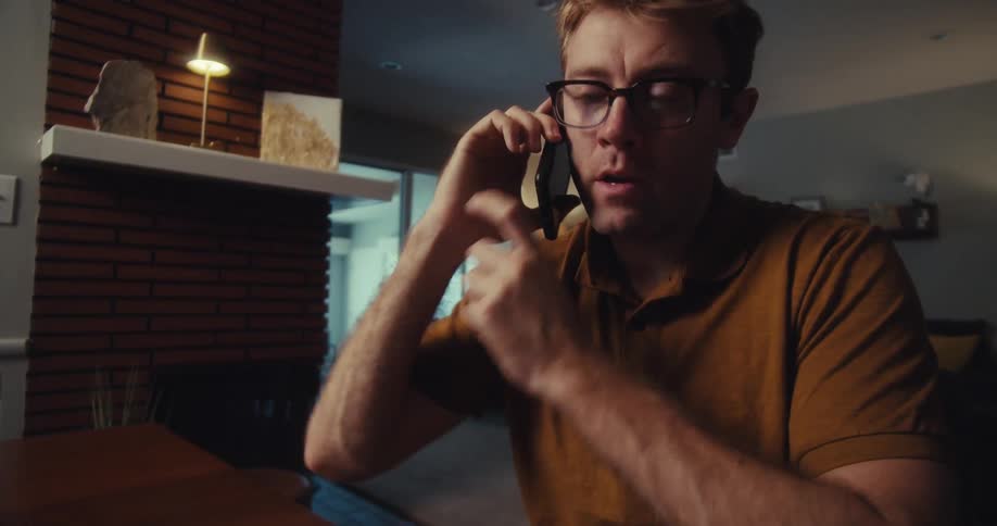 While highlighting figures on financial documents a man with glasses and a brown shirt speaks on his phone with a serious expression