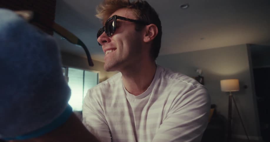 Man happily cleans his sunglasses with a cloth while singing and moving enthusiastically indoors