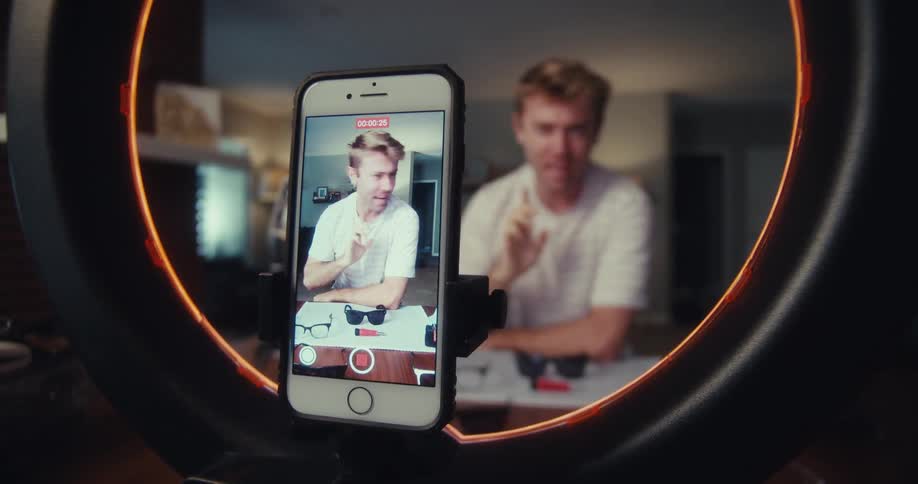 A man in a room being recorded by a cellphone mounted on a ring light