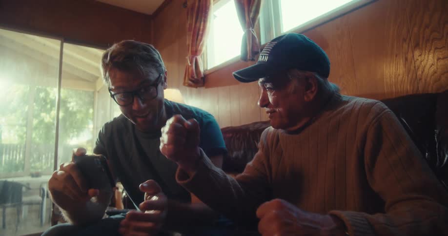 Two men are sitting on the couch looking at a phone and celebrating