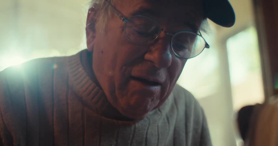 An elderly man wearing a hat and glasses holds a notebook and writes in it with a pen