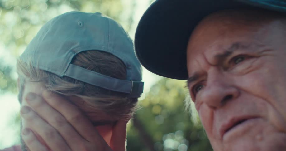 Two men wearing baseball caps one younger looking down and the other older looking up and to the side