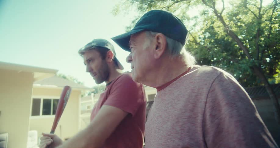 Two men hold a baseball and bat in the yard while talking