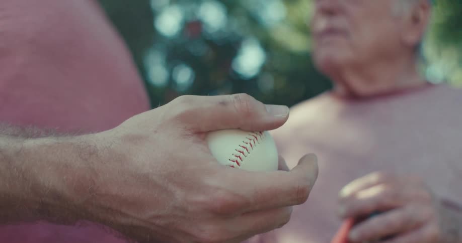 A man holding a baseball talking to an elderly man under a tree on a sunny day