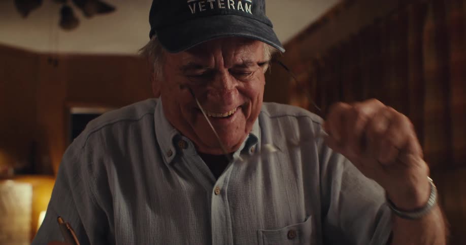 An elderly man writes at a desk then takes off his glasses and starts laughing with a coffee mug on the table beside him