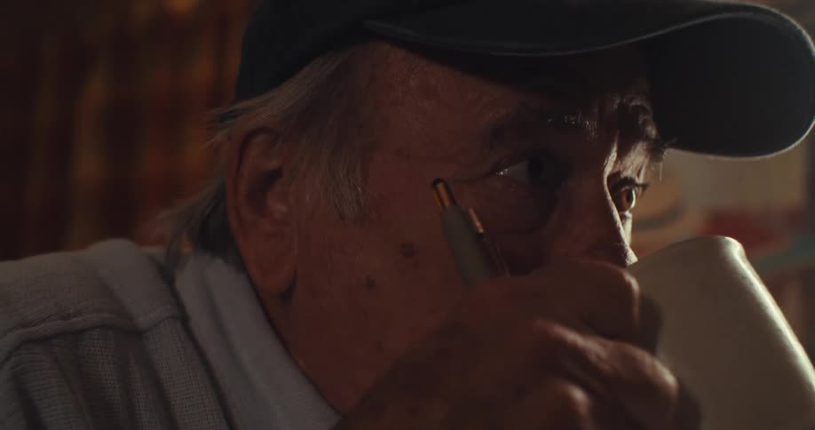 A footage of an elderly man sipping a cup of coffee while writing and also wearing his eyeglasses