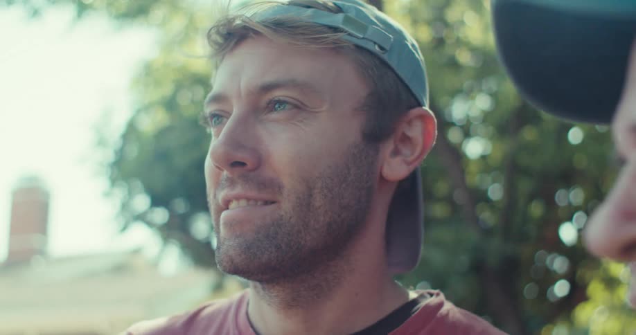 A young man and an elderly man both wearing baseball hats stand on a lawn on a sunny day engaged in conversation and smiling warmly at each other
