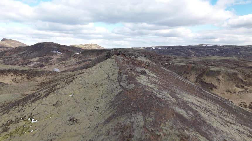 Arid mountainous landscape with sparse vegetation and small patches of snow stretches out under a partly cloudy sky