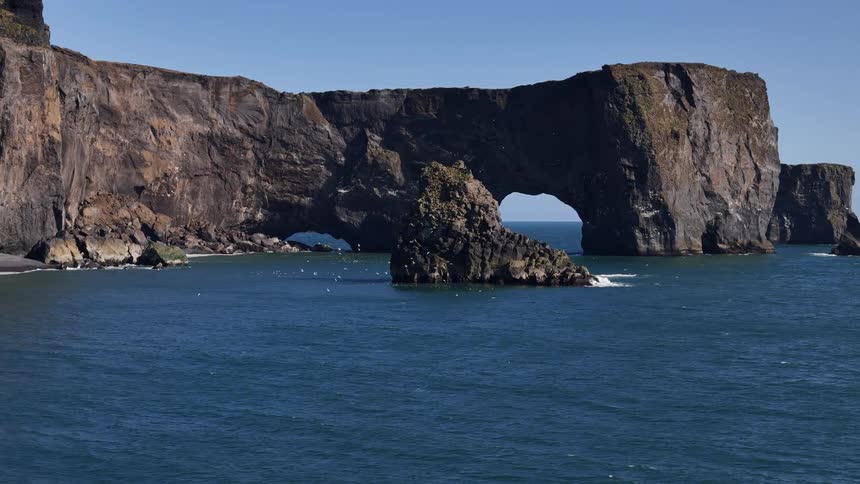 Seagulls fly over the water as waves splash against rock formations with arches in the promontory