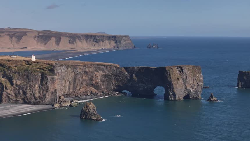 A dramatic coastal cliff with a large arch juts into the ocean under clear blue skies