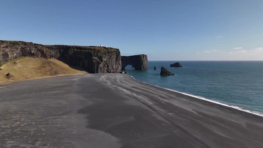 Black sand beach stretches to cliffs and rock formations in the sea under a clear blue sky