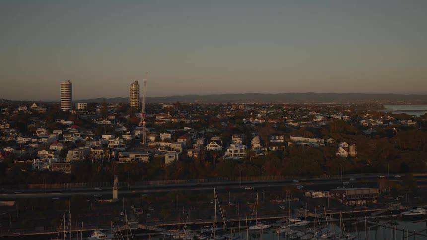 An aerial view shows a harbor filled with numerous docked sailboats bordered by a coastal urban area with houses roads and distant hills under a dusky sky