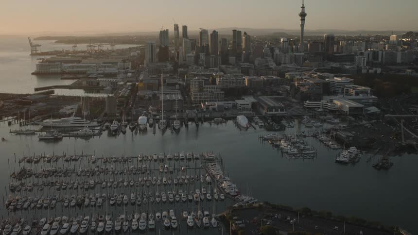 A city skyline near the shore with several sailboats docked at a dockyard as the sun rises