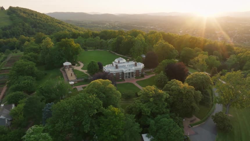 An aerial shot shows a grand mansion with a domed roof nestled amidst expansive green lawns and mature trees on a hilltop as the sun rises in the background