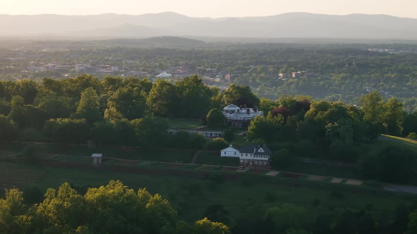 An aerial view captures a grand estate with a prominent mansion nestled on a treecovered hilltop overlooking a distant town and hazy mountain ranges