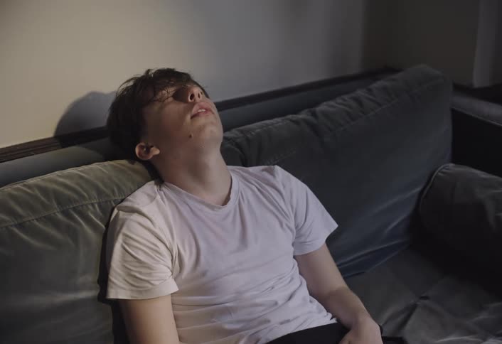 A teenage boy looks depressed and tired as he flops his head back on the couch