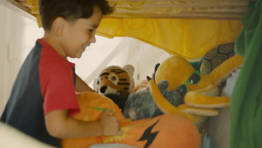 Two boys play hide and seek in a bright bedroom one searching while the other hides under a pile of stuffed animals on the bed