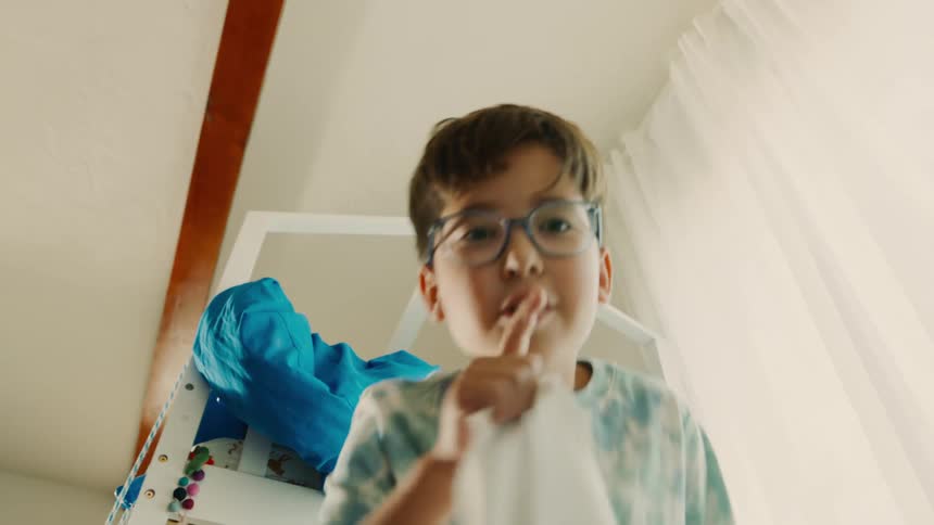 A boy playing hide and seek gestures for silence by placing a finger to his mouth then covers the camera with a blanket as if it were another player he is hiding