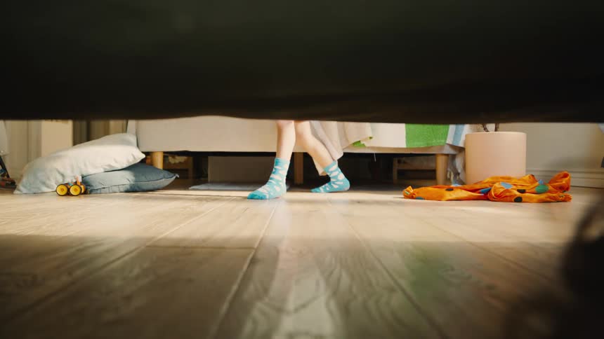 A boy wearing blue socks searches the room during a game of hide and seek looking under beds