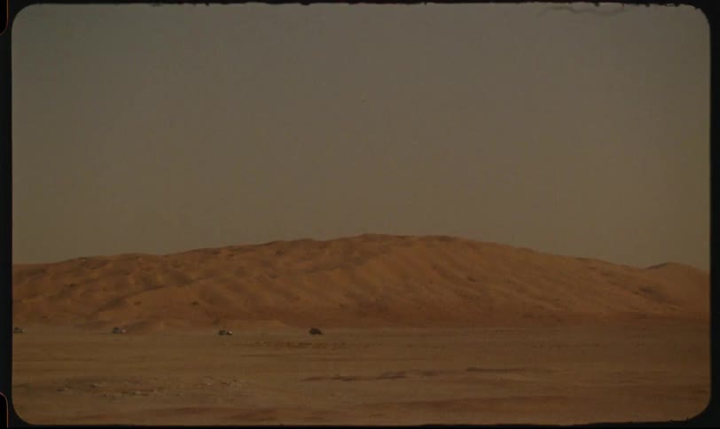 Several vehicles traverse a vast sandy landscape under a hazy sky kicking up dust as they move