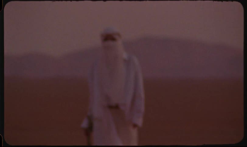 A person in desert attire stands alone in a blurry reddish desert landscape at sunset
