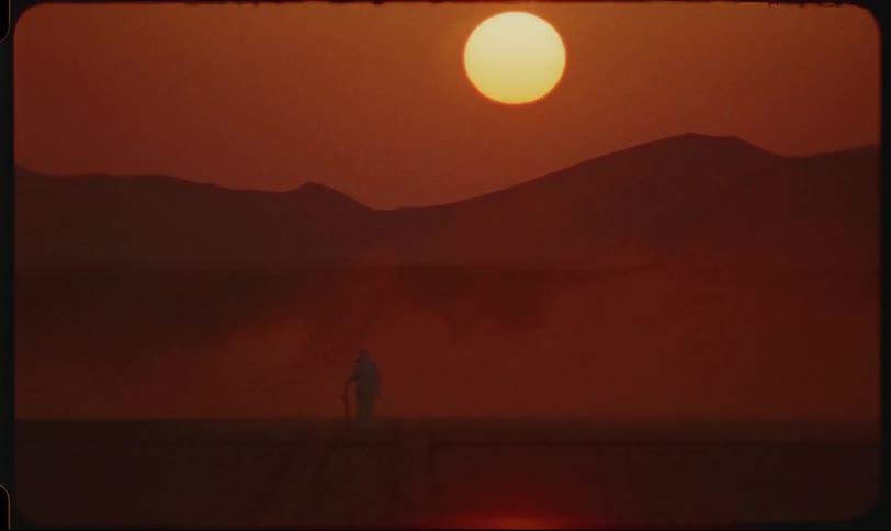 A lone person walks through a desert at sunset with large mountains silhouetted in the background
