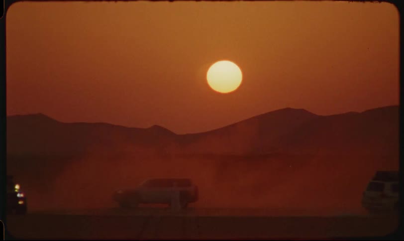 Vehicles driving in a circle with a silhouette of a person in the middle in a desert landscape with the sunset in the background