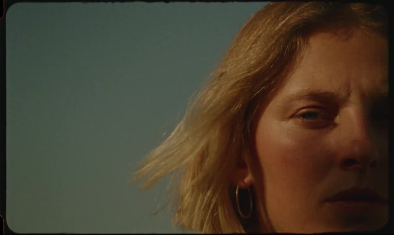A closeup of the side of a womans face with light blonde hair wearing a gold hoop earring against a clear light blue background