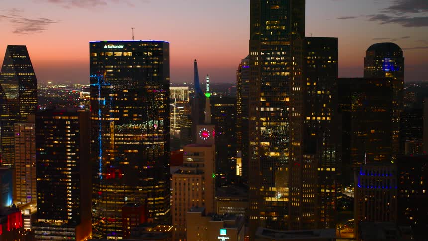 An aerial view captures the illuminated skyscrapers of the dallas city skyline against a vibrant sunset sky with numerous city lights twinkling below