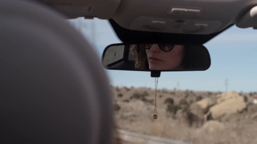 A woman drives on a desert road in the daytime and is seen in the rearview mirror