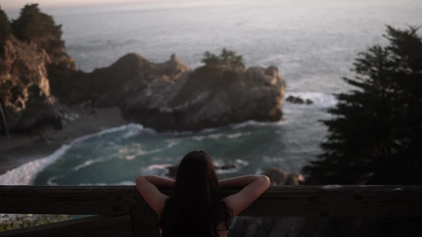 A woman looks over the railing at the ocean cove below