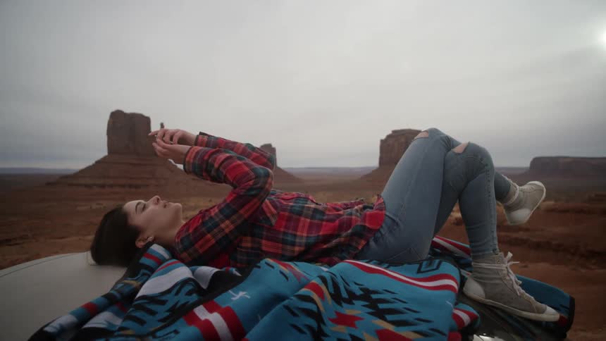 Woman lays on blanket on a car and plays with her phone