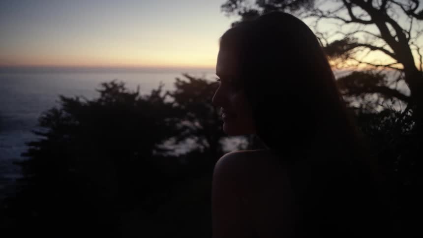A woman sits on a beach side cliff at dusk and smiles