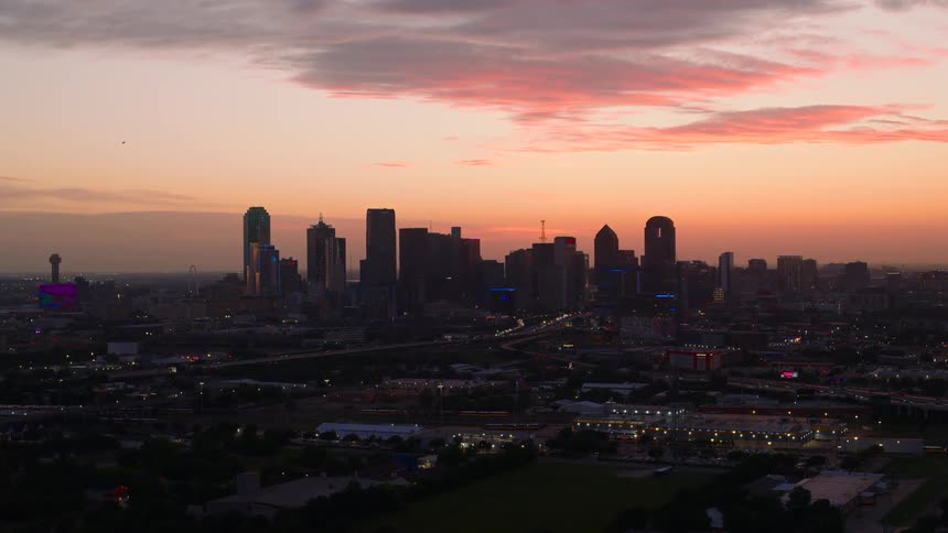 The dallas skyline is silhouetted against a vibrant colorful sunrise sky above a sprawling urban landscape with illuminated roadways and buildings