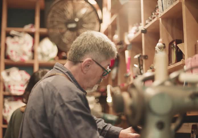 An older man is carefully working at a cluttered workstation in a dimly lit workshop