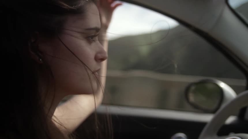 A woman drives with the window down while her hair blows around her face