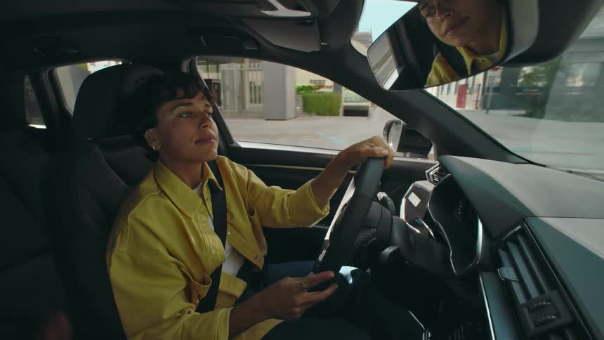 A woman with short dark hair and a yellow jacket, with her reflection in the rearview mirror, drives a car through a city on a sunny day