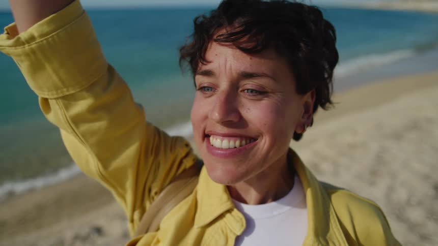 A woman in a yellow shirt walking in the sand along the beach, looks ahead and furrows her brows as she sees someone in the distance and then waves when she recognizes them.