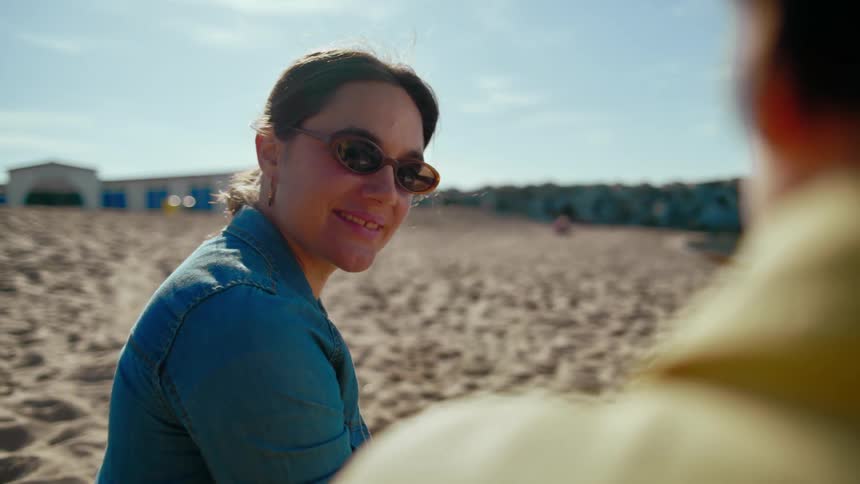 Two women sitting on the beach sand at midday chatting and laughing together.
