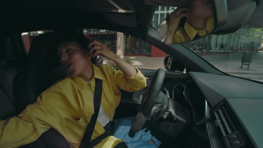 A woman is driving in her car and using her hairbrush as a microphone while she sings to her reflection in the rearview mirror and gestures dramatically to the music.