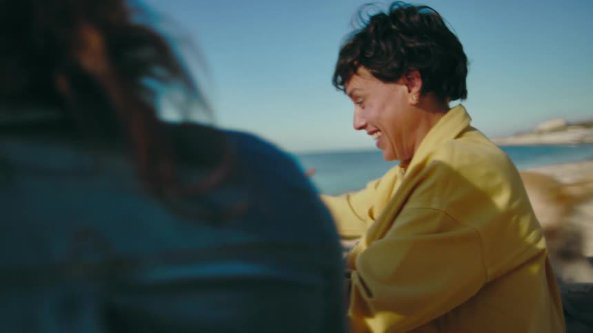 Two women sitting in the sand at the beach next to the crystal blue Mediterranean Sea talking excitedly as their dogs play on a sunny day