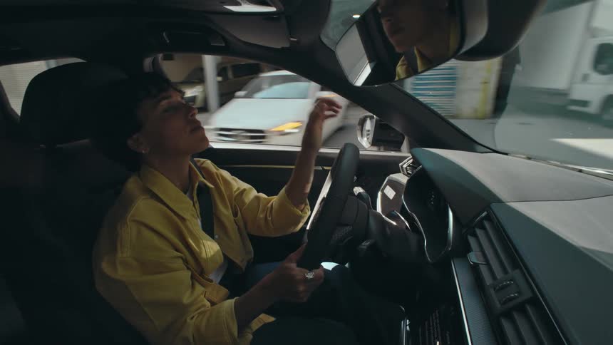 Excited woman driving a car along city streets as she smiles, bobs her head, and drums the steering wheel with her hand to the beat of music.