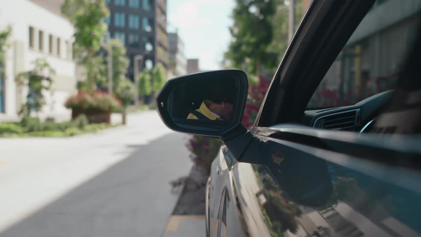 POV looking at the side rearview mirror of a car as a female driver closes the door and puts on her seatbelt.