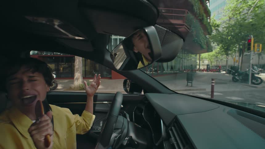 A woman in a yellow shirt dramatically sings into a hairbrush that she is pretending is a microphone, while sitting in the drivers seat of her car as she waits at a stoplight.