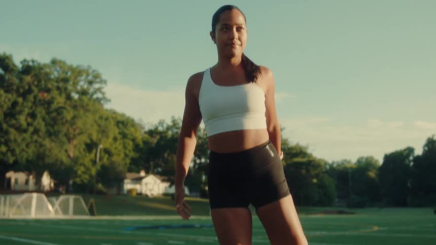 A woman with dark hair pulled back in a ponytail stretches her legs on a green athletic field marked with white lines